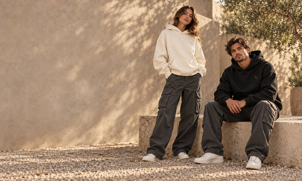Two models in bone and black raven hoodies with charcoal cargos in a sunlit stone courtyard