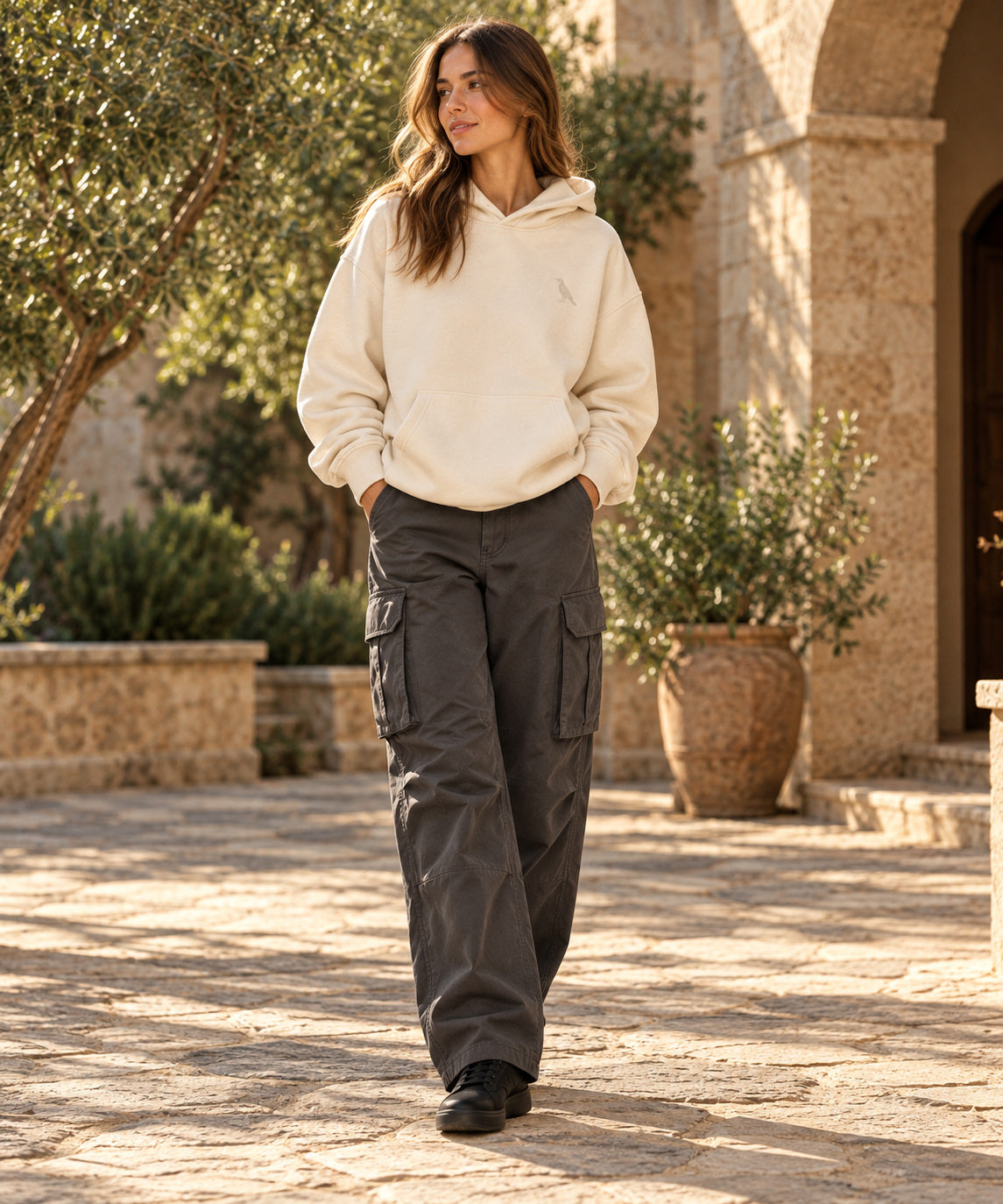 Woman in a bone raven hoodie and charcoal cargo pants in a sunlit stone courtyard